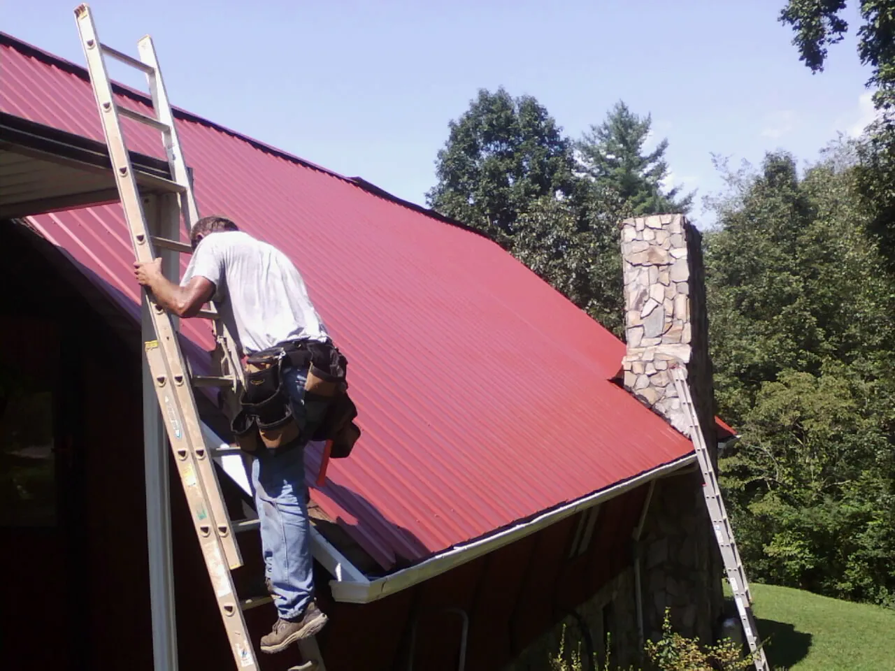 Roofer on a ladder beside a red metal roof and stone chimney — Soffit & Fascia Repair by Roofing Brothers in Whitmore